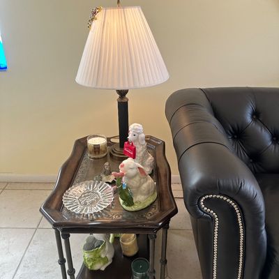 Sale Photo Thumbnail #2: Vintage dark wood two-tier end table topped with a pleated-shade lamp, ceramic figurines including a bunny and poodle, glass dish, and candles.