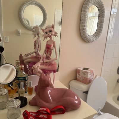 Sale Photo Thumbnail #65: Eclectic bathroom vanity decor featuring a pink ceramic reclining cat statue, an anthropomorphic ballerina cat figure, and vanity bottles.