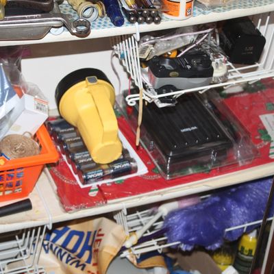 Sale Photo Thumbnail #134: Storage cabinet filled with household supplies, including a yellow lantern flashlight, batteries, electronics, hand tools, and a purple duster.