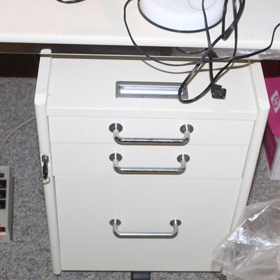 Sale Photo Thumbnail #90: White metal three-drawer mobile filing pedestal with chrome handles positioned underneath a matching desk, shown alongside a power strip.
