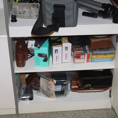 Sale Photo Thumbnail #82: Collection of camera equipment and electronics inside a white cabinet, including a Logitech webcam, Canon PowerShot boxes, camera bags, and a tripod.
