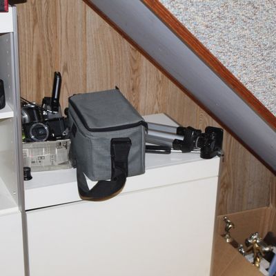 Sale Photo Thumbnail #77: Assorted camera equipment including a gray padded shoulder bag, compact digital camera, and tripod, set alongside vintage binoculars on a shelf and a box of trophies.