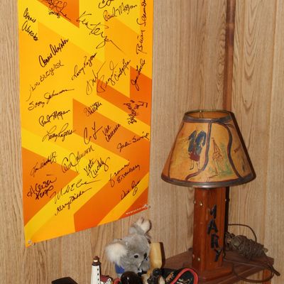 Sale Photo Thumbnail #116: Autographed Olympic-themed poster hanging beside a personalized wooden table lamp and assorted small collectibles on a wall shelf.