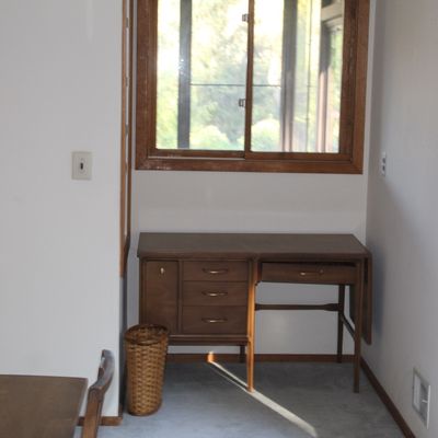Sale Photo Thumbnail #25: Mid-century modern walnut desk featuring brass pulls and a drop-leaf side, positioned beneath a wooden-framed window.