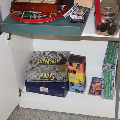 Sale Photo Thumbnail #80: Collection of vintage board games and playing accessories, including Moonshot, The Sierra Club Game, and a metal dominoes tin, stored in a white cabinet.