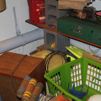 Sale Photo Thumbnail #128: Basement storage unit filled with mixed items including a vintage wood cabinet speaker, croquet mallets, a green metal tackle box, and a red Gloy Heat-Pal heater.