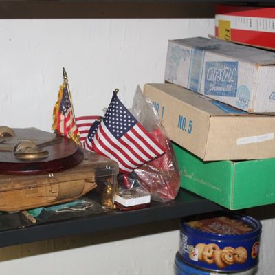 Sale Photo Thumbnail #106: Collection of shelf items including small American flags, a Masonic wooden plaque, vintage boxed glassware, and cookie tins.