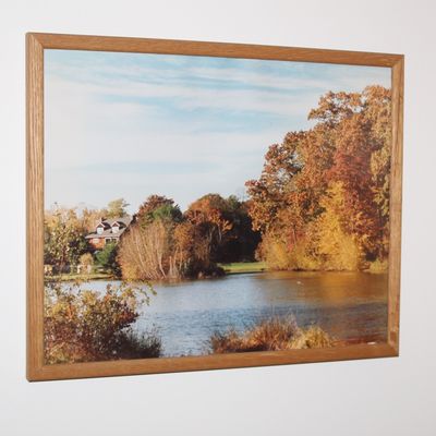 Sale Photo Thumbnail #4: Framed photographic print depicting an autumn landscape scene with a house near a lake, housed in a simple light oak wooden frame.