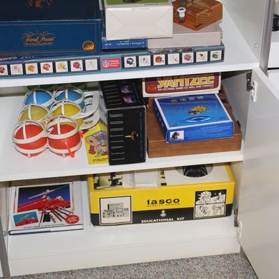 Sale Photo Thumbnail #81: Collection of vintage board games, science novelties, and educational kits stored inside a white cabinet, including Trivial Pursuit, Levitron, and a Tasco microscope set.