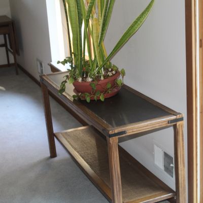 Sale Photo Thumbnail #24: Mid-century wooden console table with a lower woven shelf and decorative corner brackets, featuring a potted snake plant on top.