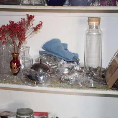Sale Photo Thumbnail #43: Cabinet shelf containing a collection of glassware, silver-plated items wrapped in plastic, a red Bohemian-style miniature vase with dried flowers, and a corked glass bottle.