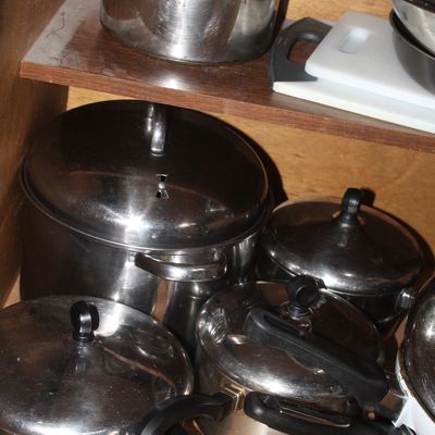 Sale Photo Thumbnail #70: Collection of stainless steel pots and pans with black handles, stored inside a wooden kitchen cabinet alongside a plastic cutting board.