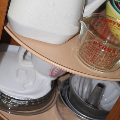 Sale Photo Thumbnail #69: Corner cabinet Lazy Susan filled with kitchenware, including a vintage Pyrex 4-cup glass measuring cup, a speckled plastic watering pitcher, and metal cake pans.
