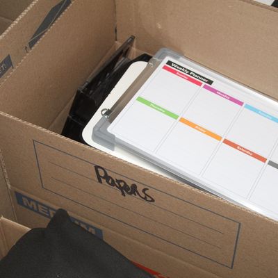Sale Photo Thumbnail #144: Dry-erase weekly planner whiteboard with colorful magnets resting inside a moving box packed with office supplies.