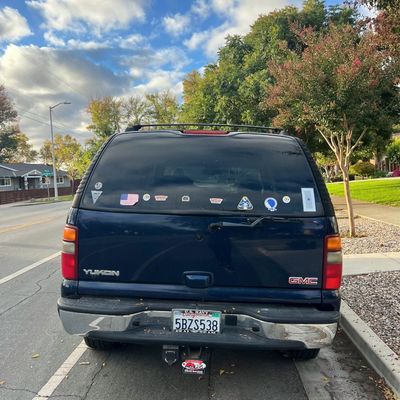 Sale Photo Thumbnail #36: Rear view of a dark blue GMC Yukon SUV parked along a street curb, featuring bumper stickers and military decals on the rear window.