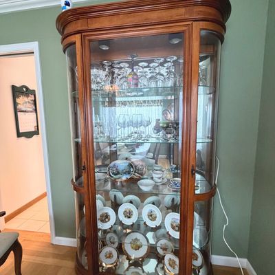 Sale Photo Thumbnail #57: Traditional wooden curio display cabinet with curved glass sides and interior lighting. Filled with fine stemware, teacups, and decorative collector plates.