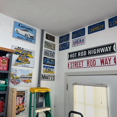 Sale Photo Thumbnail #40: Collection of vintage-style automotive tin signs and various state license plates mounted on garage walls above a shelving unit and door.
