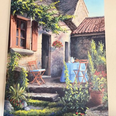 Sale Photo Thumbnail #14: Framed canvas print depicting a sunlit European courtyard scene with a table set for tea, lush greenery, and potted flowers, signed in the lower right corner.