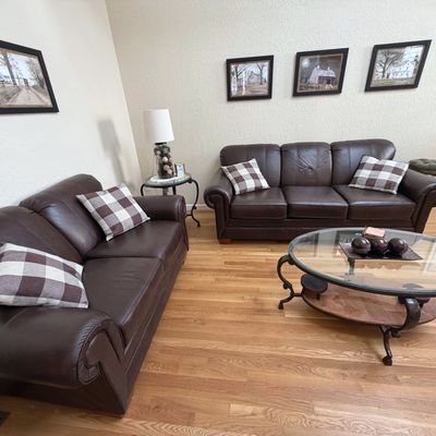 Sale Photo Thumbnail #49: Living room set including a dark brown leather sofa and matching loveseat with plaid accent pillows, alongside an oval glass-top coffee table.