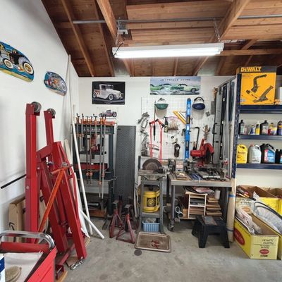 Sale Photo Thumbnail #44: Workshop garage filled with automotive and woodworking tools, including a red hydraulic shop crane, hydraulic press, bench grinder, pegboard organization, and shelving units with automotive fluids.
