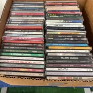 Cardboard box filled with assorted compact discs in jewel cases, including various pop, rock, and holiday titles.