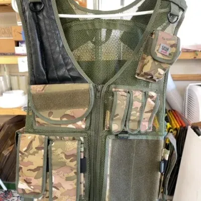 Olive green mesh tactical shooting vest with camouflage pockets and a black padded right shoulder guard.