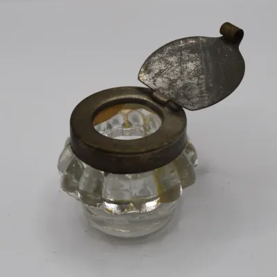 Sale Photo Thumbnail #62: Antique molded clear glass inkwell featuring a hinged metal flip-lid with an aged patina.