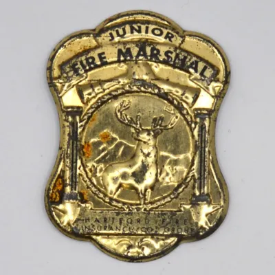 Sale Photo Thumbnail #37: Vintage Junior Fire Marshal tin pin badge, issued by the Hartford Fire Insurance Company Group, featuring embossed lettering and the signature stag logo with some surface wear and patina.