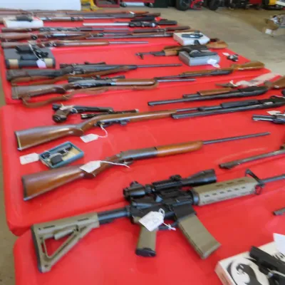 Sale Photo Thumbnail #3: Large collection of rifles, shotguns, and handguns arranged on red tables, including modern sporting rifles, lever-action rifles, and various pistols.