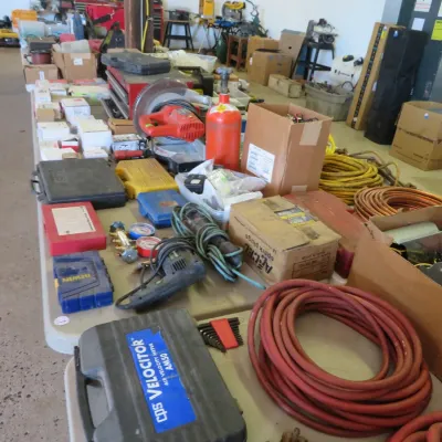 Sale Photo Thumbnail #27: Workshop and electrical supplies on folding tables, including a CPS Velocitor air velocity meter case, coiled hoses and copper tubing, power tools, and various boxed hardware.