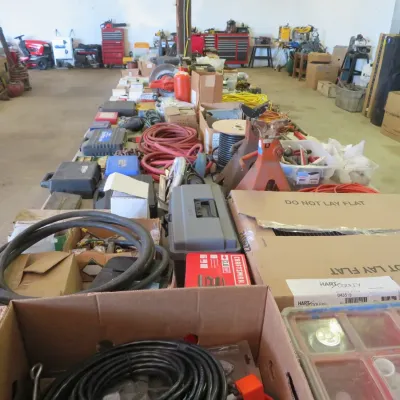 Sale Photo Thumbnail #26: Large collection of workshop and garage items displayed on long folding tables, including extension cords, jack stands, power tool cases, and boxed hardware.