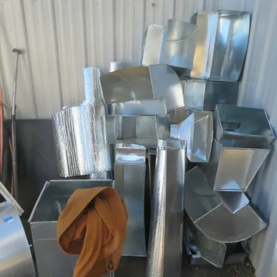 Sale Photo Thumbnail #47: Assorted galvanized sheet metal HVAC ductwork components, elbows, transitions, and foil duct insulation stacked in a workshop corner.