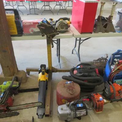 Sale Photo Thumbnail #41: Collection of outdoor power equipment, power tools, and shop accessories, including chainsaws, a DeWalt leaf blower, shop vacuum, and jack stands arranged on foldable tables and the floor.