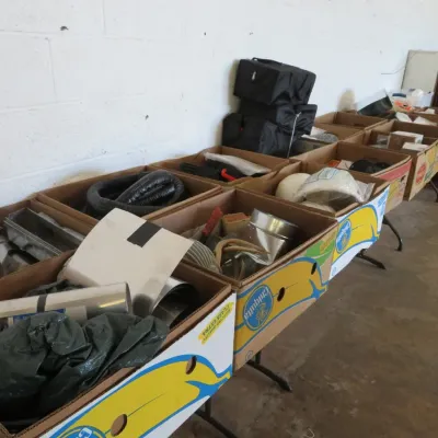 Sale Photo Thumbnail #60: Assorted cardboard banana boxes filled with miscellaneous hardware, ducting, equipment parts, and household items, arranged on folding tables.