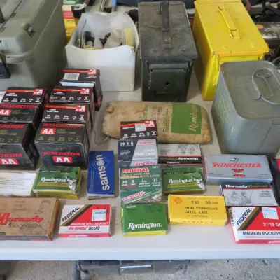 Sale Photo Thumbnail #53: Assortment of ammunition boxes and metal military-style ammo cans displayed on a folding table, featuring brands like Winchester, Remington, Federal, and Hornady.