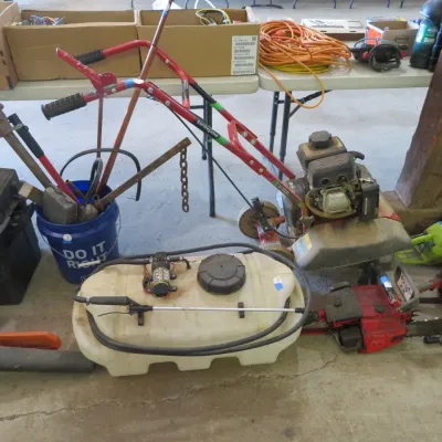 Sale Photo Thumbnail #40: Collection of outdoor power equipment and tools, including a red gas-powered tiller, a tow-behind tank sprayer, chainsaws, and hand tools.