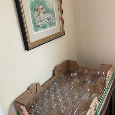 Sale Photo Thumbnail #142: Cardboard crate filled with assorted clear glassware, including wine glasses and champagne flutes, displayed below a framed cheetah print.
