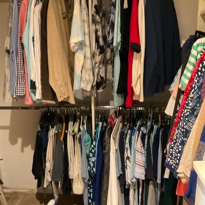 Sale Photo Thumbnail #171: A collection of men's and women's clothing hanging in a closet, including button-down shirts, jackets, and tops, alongside a portable steamer on the floor.