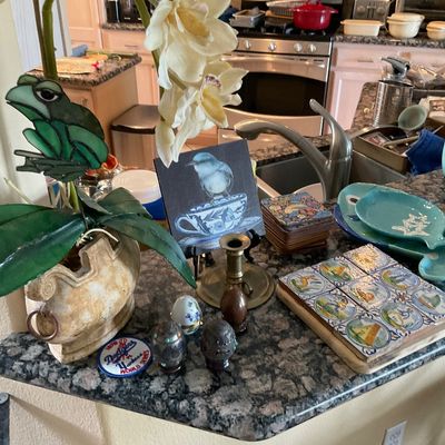 Sale Photo Thumbnail #97: Collection of decorative household items arranged on a kitchen counter, including a potted faux orchid with a stained glass frog insert, small bird painting, ceramic tile trivet, brass candlestick, and decorative eggs.