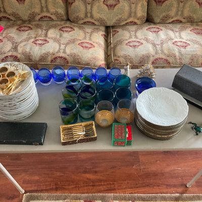 Sale Photo Thumbnail #78: Collection of tabletop items including cobalt blue glassware, stacks of ceramic dinner plates, boxed flatware, and decorative accents arranged on a folding table in front of a patterned sofa.