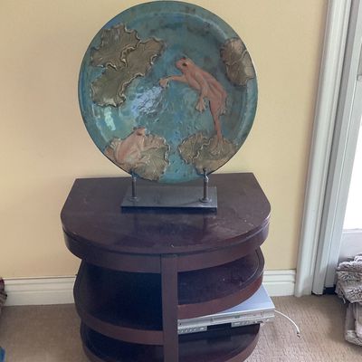 Sale Photo Thumbnail #140: Large glazed ceramic charger featuring relief frogs and lily pads, displayed on a metal stand atop a dark wood three-tiered accent table.