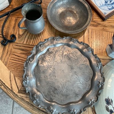 Sale Photo Thumbnail #126: Vintage pewter service pieces including a large engraved round tray, a small pitcher, and a hammered bowl arranged on an oak parquet surface.