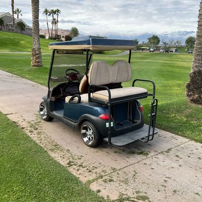 Sale Photo Thumbnail #6: Dark blue four-passenger golf cart with a rear-facing fold-down bench seat, custom chrome wheels, and a hardtop canopy, parked outdoors on a paved pathway.