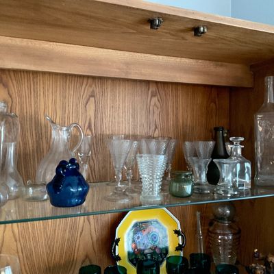 Sale Photo Thumbnail #130: Collection of vintage glass stemware, bottles, decanters, and decorative glassware displayed on the glass shelf of an oak hutch.