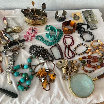 Sale Photo Thumbnail #113: Eclectic assortment of vintage fashion jewelry, wristwatches, magnifying glasses, and small collectibles, including turquoise, amber, and beaded necklaces displayed on a white cloth surface.