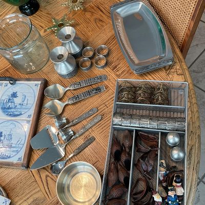 Sale Photo Thumbnail #122: Collection of vintage tabletop items including metal serving utensils, pewter-style candlesticks, a wire cutlery organizer filled with carved wooden and metal napkin rings, and ceramic tile trivets on a round oak table.