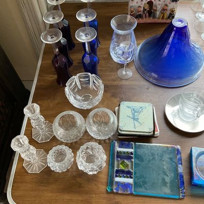 Sale Photo Thumbnail #144: Collection of glass and tableware featuring cobalt blue and purple wine glasses, clear cut-crystal bowls and candleholders, decorative coasters, fused glass trays, and a ceramic village scene figurine.