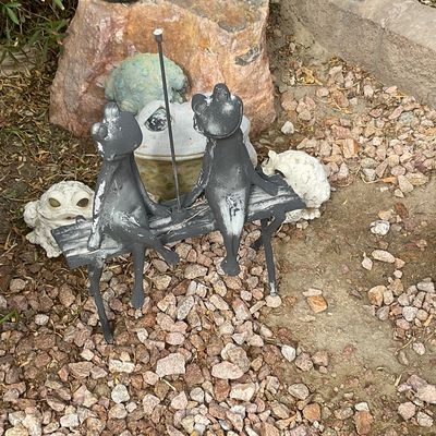 Sale Photo Thumbnail #34: Cast metal garden sculpture of two frogs sitting on a bench, displayed alongside weathered stone frog statues in a gravel landscape.