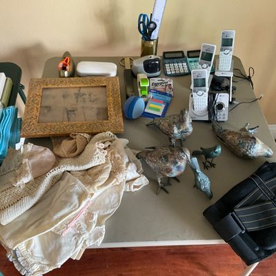 Sale Photo Thumbnail #71: Folding table displaying a collection of household goods, including bronze bird figurines, cordless AT&T phones, office supplies, vintage textiles, and a framed photo.