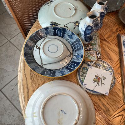 Sale Photo Thumbnail #121: Collection of antique hand-painted ceramic plates, decorative tiles, and Delftware blue and white vases displayed on a wooden table, showing visible age, wear, and previous repairs.